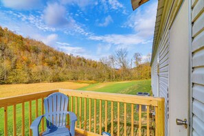 A wooden deck with a blue Adirondack chair, a view of a grassy field, and a forested hillside.