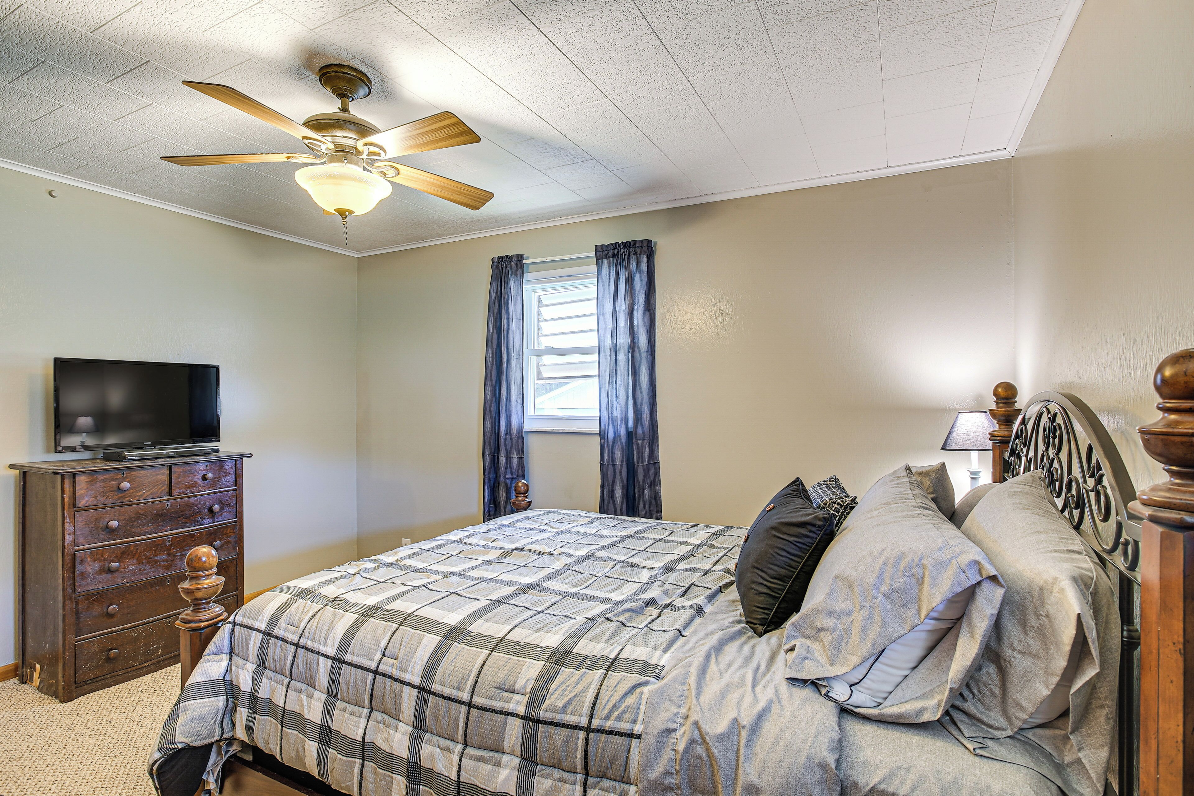 A bedroom with a bed, a ceiling fan, a dresser, a television, and a window with curtains.