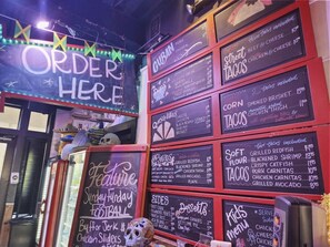 Un panneau affichant le menu avec différents plats et leurs prix, un panneau néon indiquant « COMMANDES ICI » et un panneau annonçant le « Festival spécial du dimanche ».