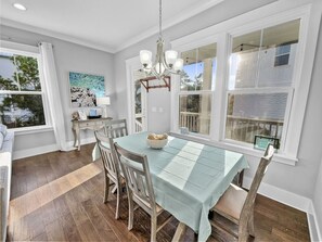 A dining area with a table set for four, surrounded by wooden chairs, a chandelier, and a view of the outdoors through large windows.