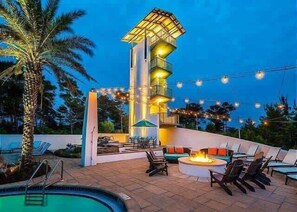 Un espace aménagé au bord de la piscine, avec un foyer, des chaises longues et un grand immeuble doté de nombreux balcons.