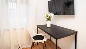 Een kleine, moderne kamer met een houten vloer, een witte stoel, een donkerhouten tafel, een witte vaas met bloemen en een grote flatscreen televisie aan de muur.