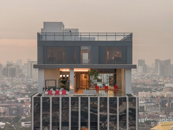 A modern two-story building with a glass facade, a rooftop terrace with red chairs, and a cityscape view.