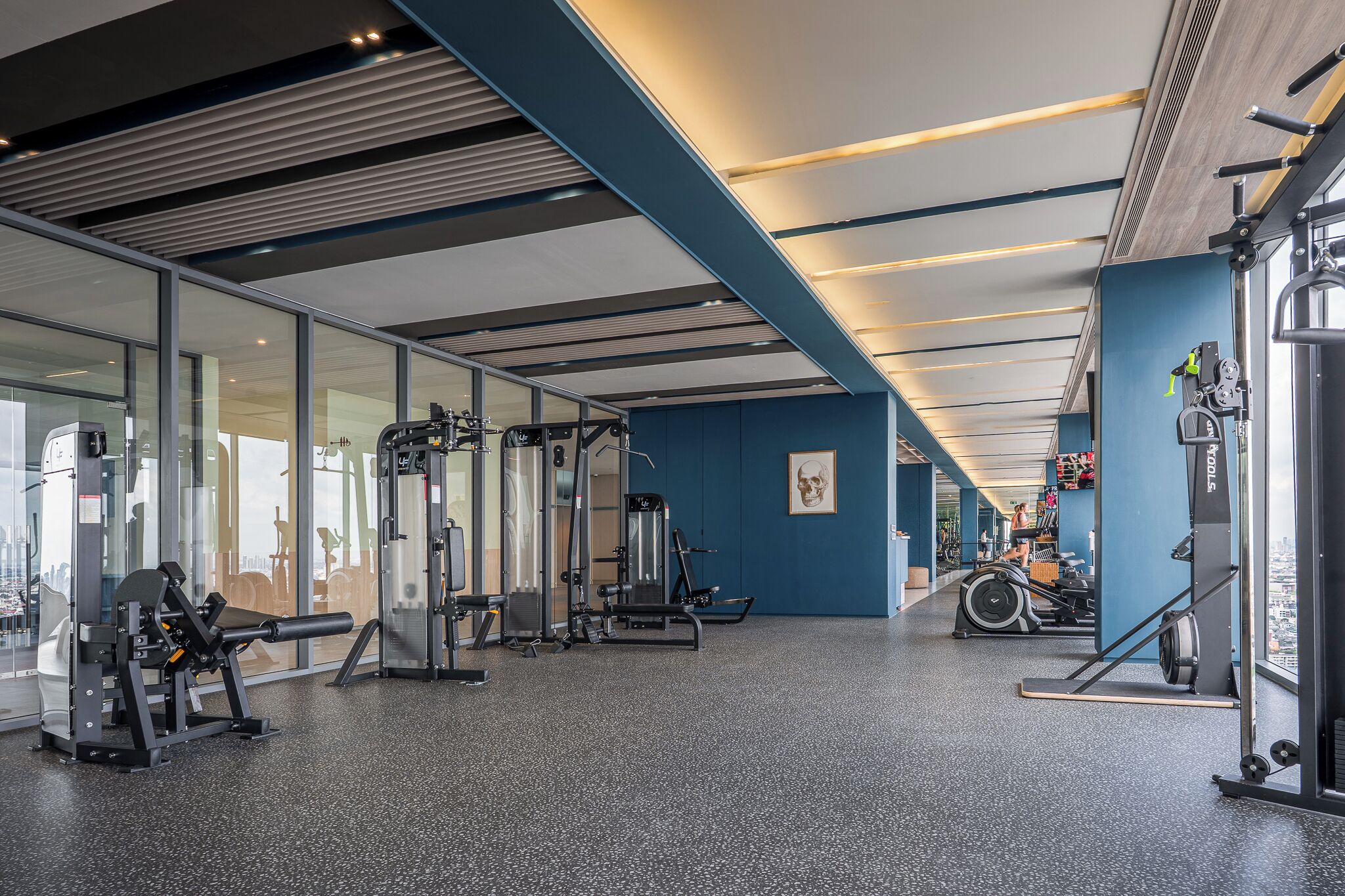A gym with various exercise machines, glass walls, and a blue accent wall.
