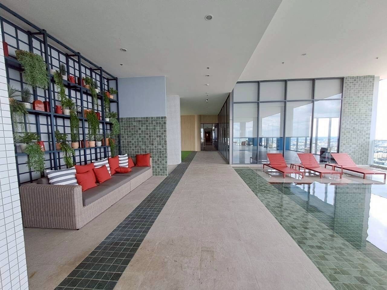 Een moderne binnenruimte met een lange gang, een zithoek met rode kussens, een wand met potplanten en een glazen balustrade met uitzicht op een stadsbeeld.