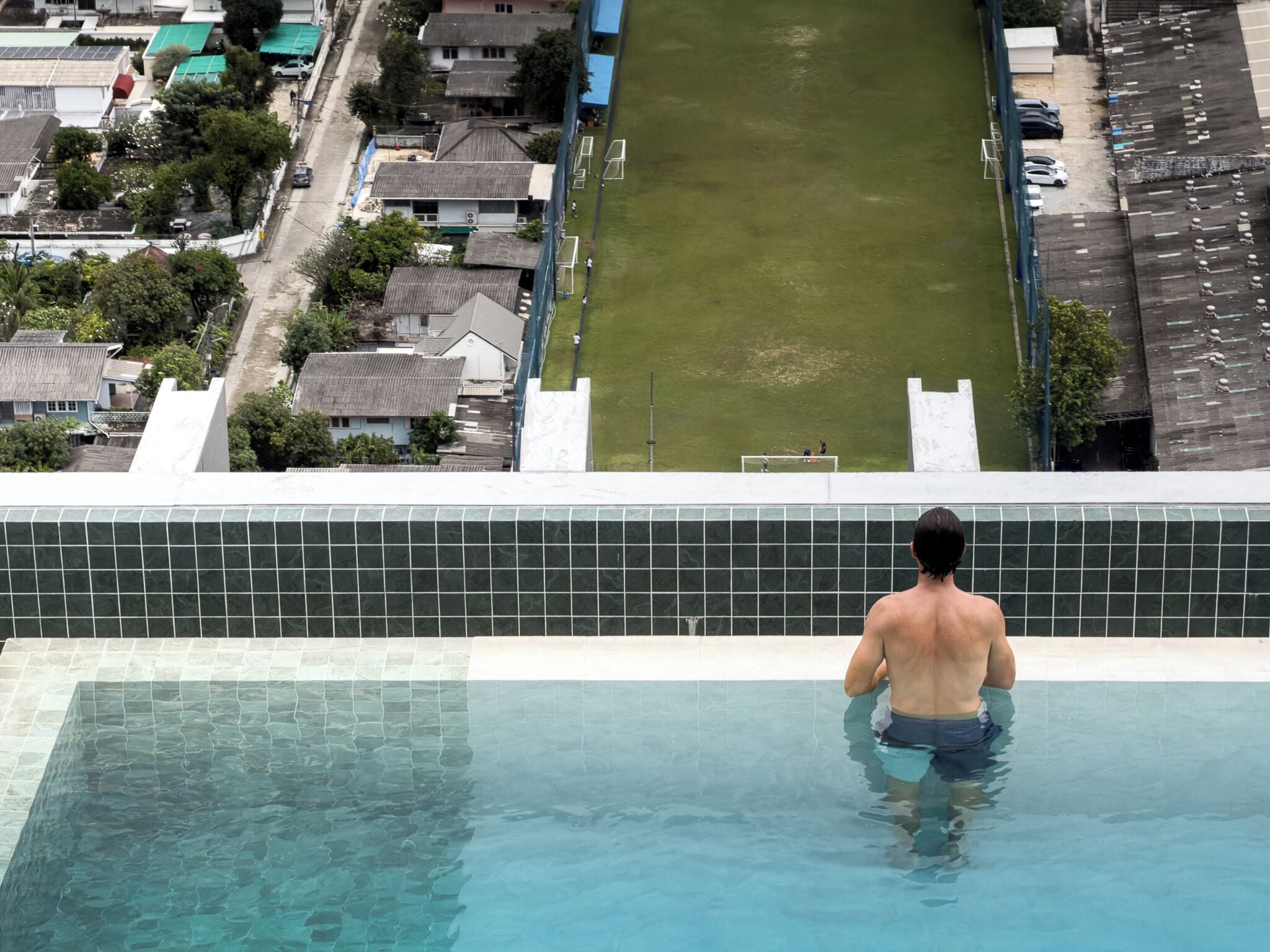 Un homme se tient au bord d’une piscine, surplombant un quartier résidentiel avec des maisons et un parking.