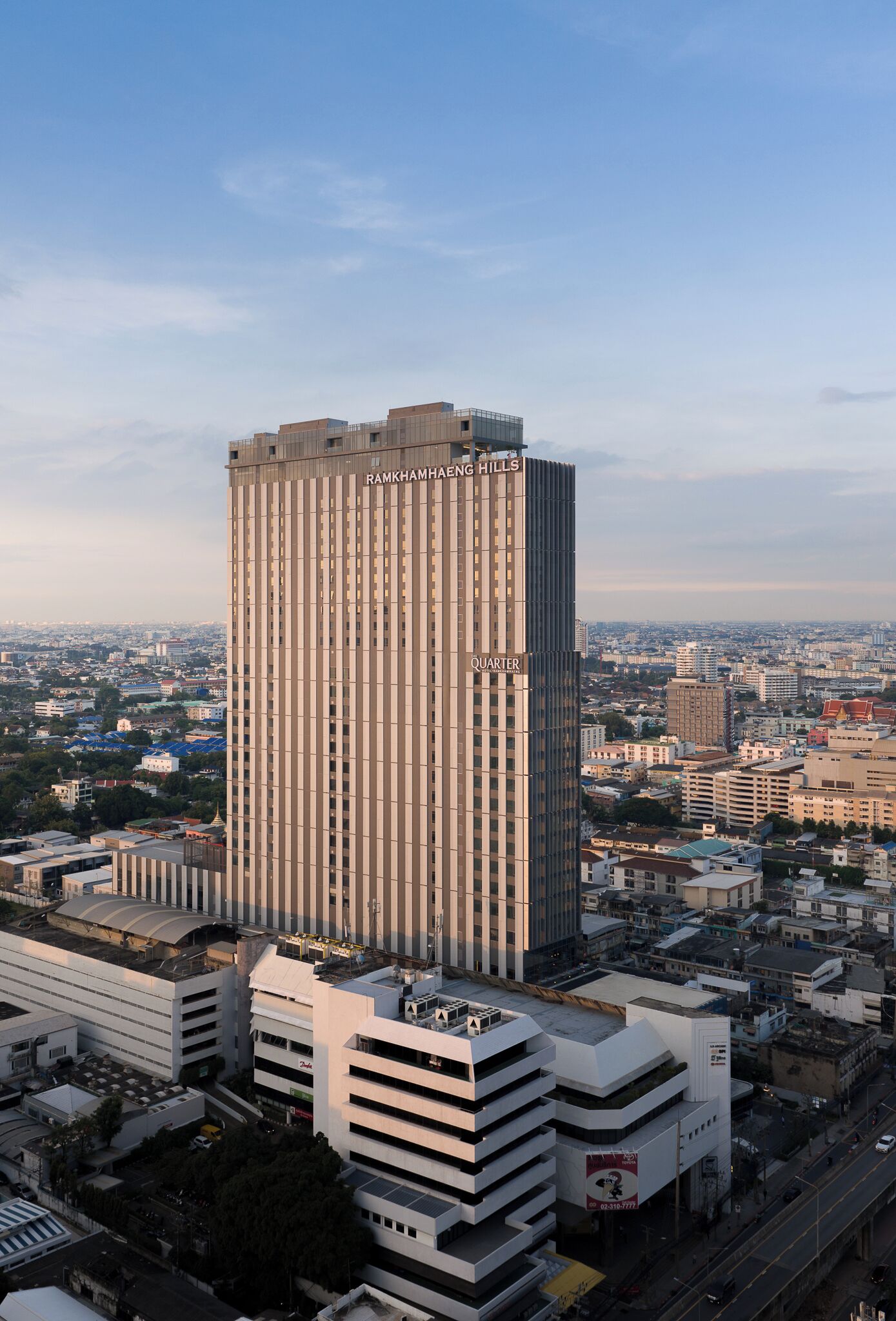 Un palazzo alto con la scritta "RAMKHAMHENG HILLS QUARTER" sulla facciata, circondato da altri edifici e un cielo sereno.