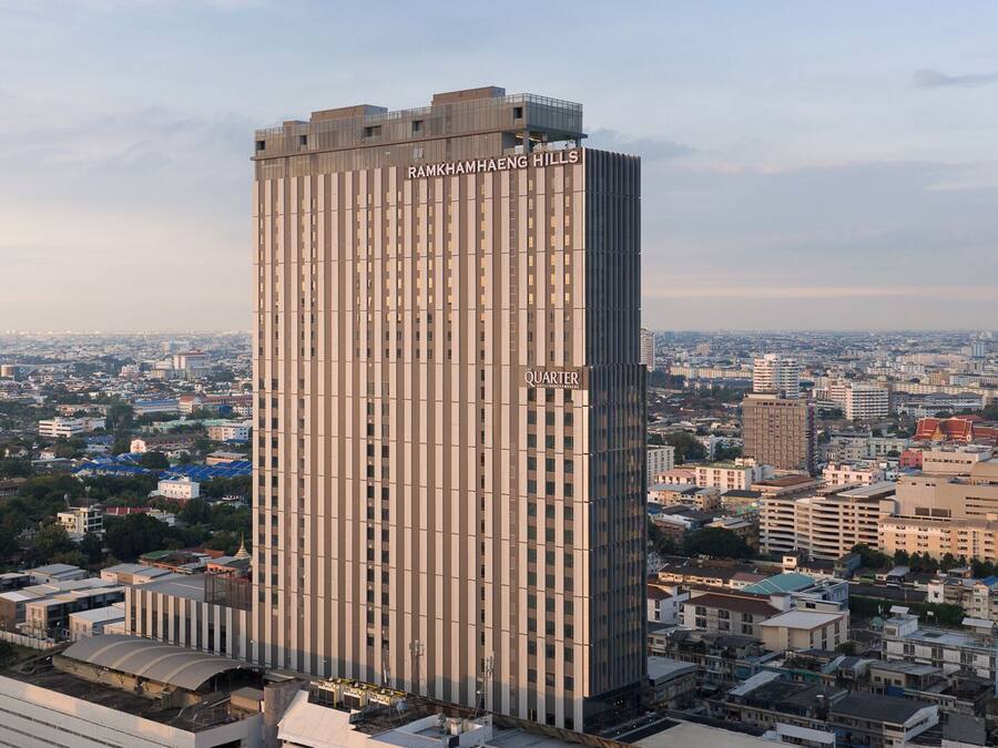 Un palazzo alto con la scritta "RAMKHAMHENG HILLS QUARTER" sulla facciata, circondato da altri edifici e un cielo sereno.
