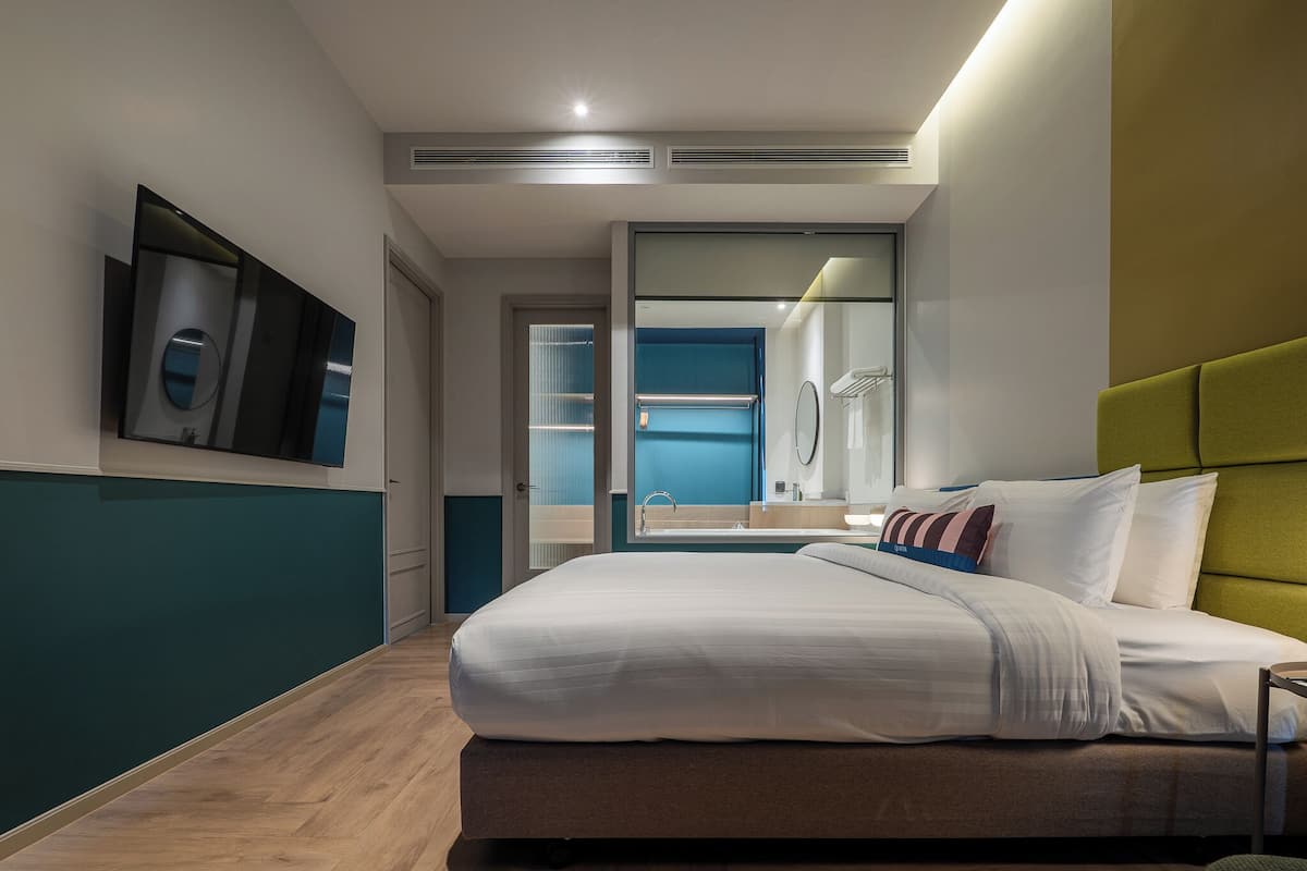 Une chambre d’hôtel moderne avec un grand lit, un miroir, une tête de lit recouverte d’un tissu vert et une vue sur la salle de bain.