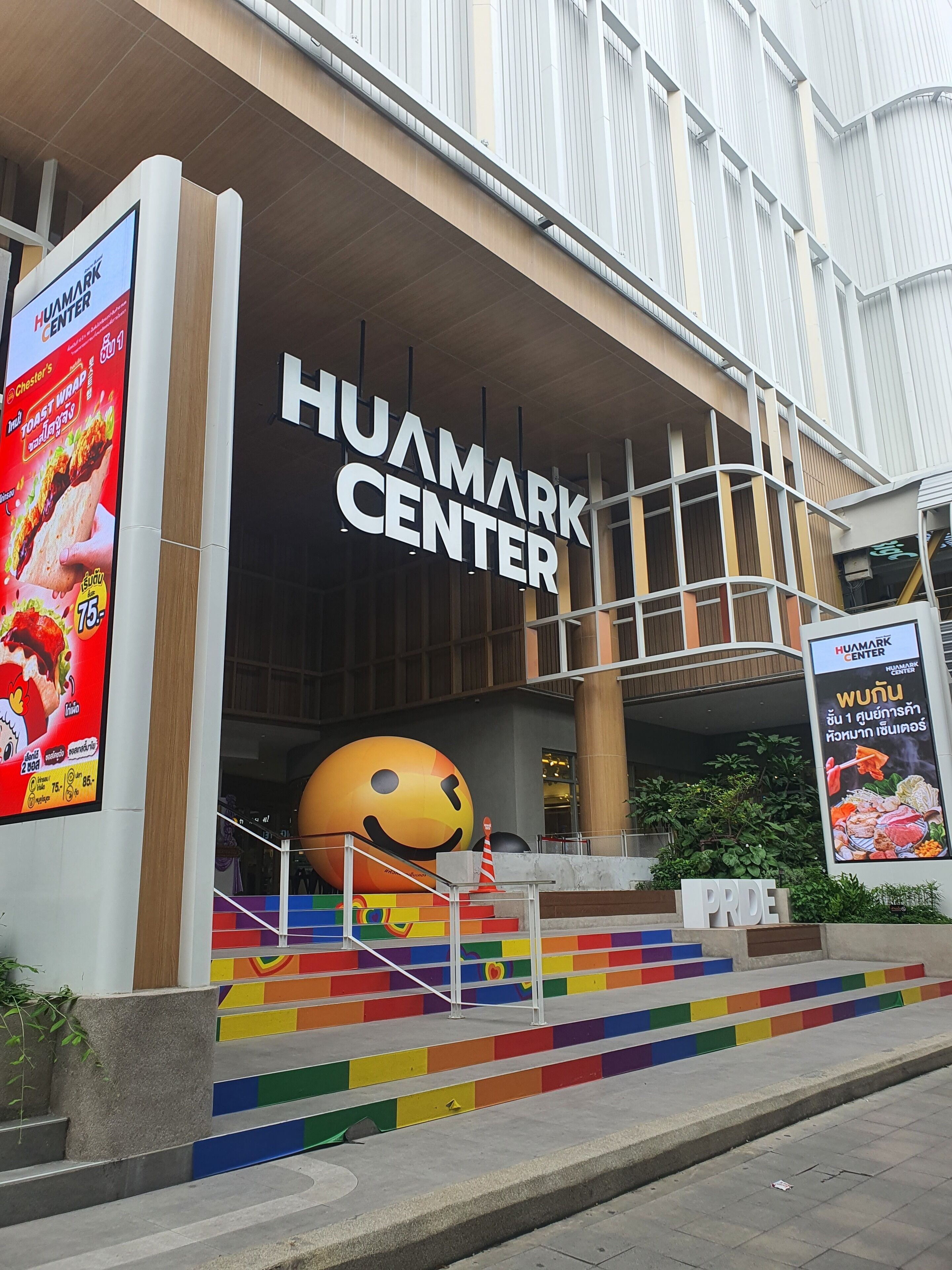 The exterior of the Huamark Center with a large smiley face sculpture at the entrance, colorful steps leading up to the doors, and various advertisements on the walls.