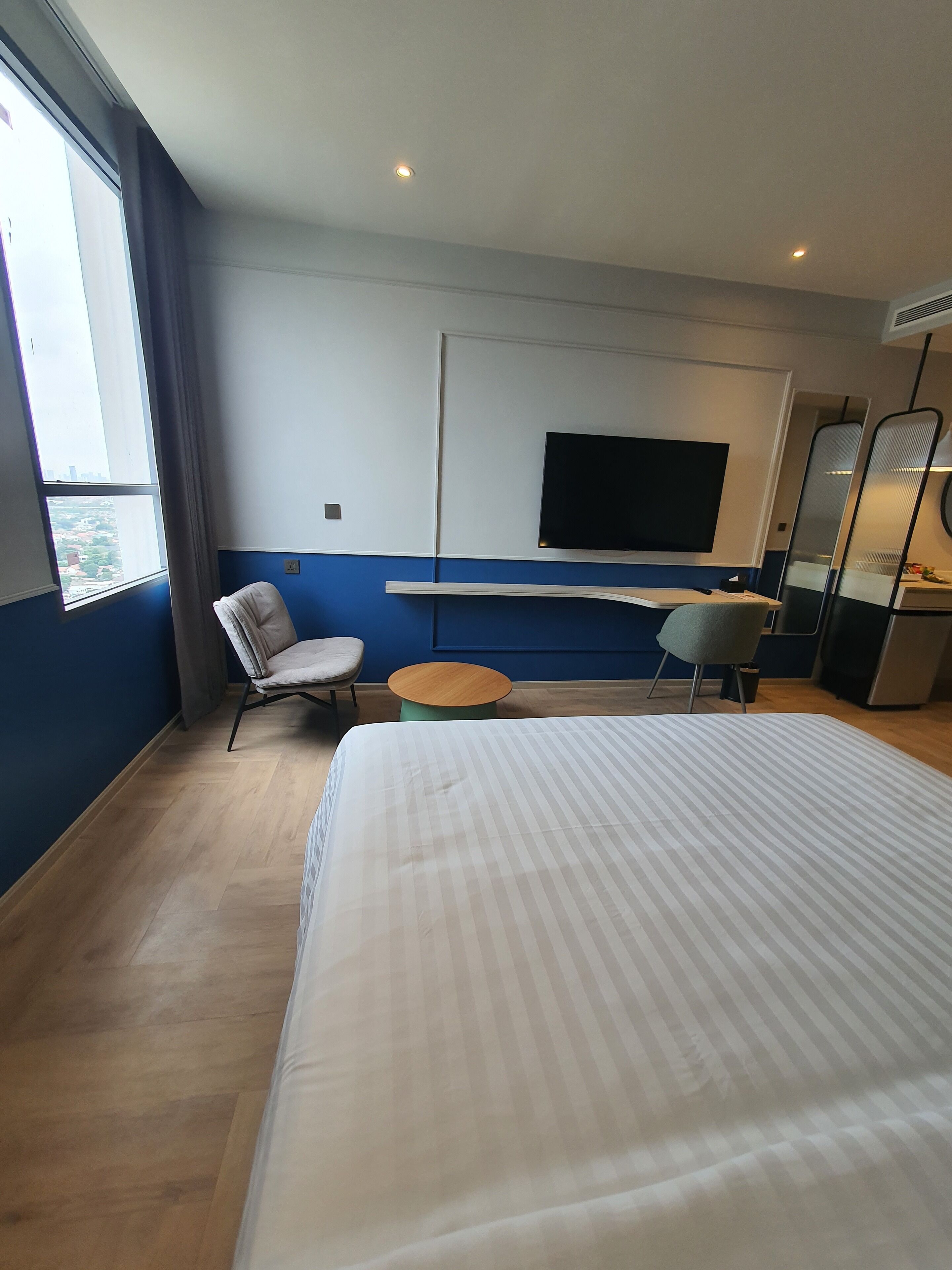 A hotel room with a large bed, a desk, a chair, a small round table, a TV on the wall, and a window with curtains.