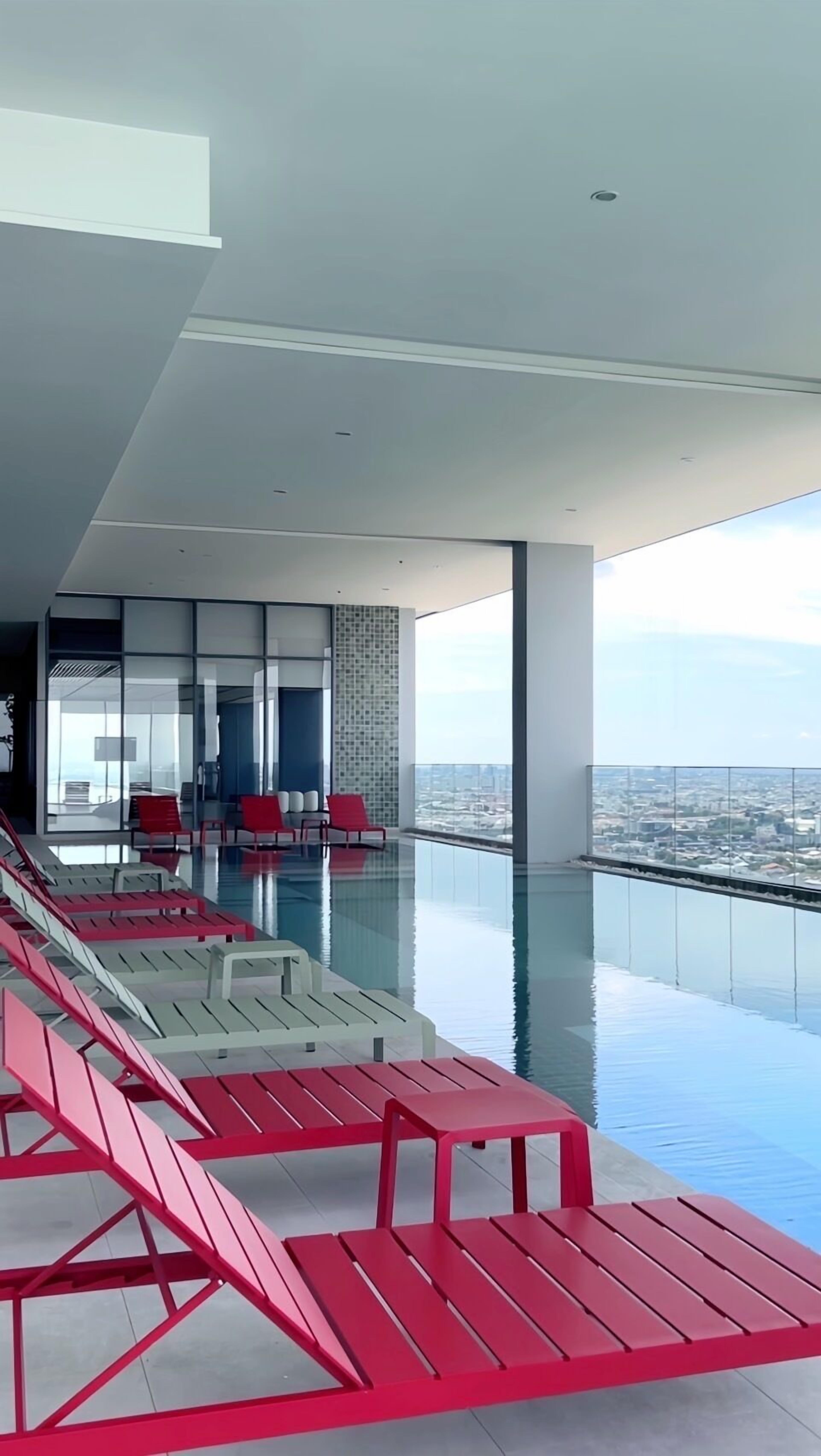 A modern infinity pool with red and white lounge chairs, a city view, and a spacious interior with a high ceiling.