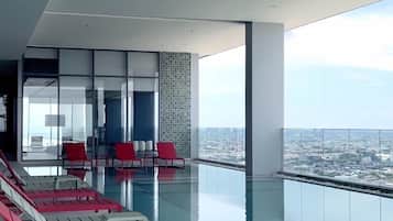 A modern infinity pool with red and white lounge chairs, a city view, and a spacious interior with a high ceiling.
