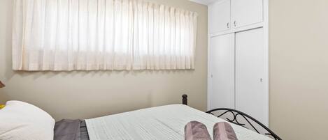 A neatly made bed with a white comforter and a rolled-up towel, a white pillow, and a beige wall with a window covered by sheer curtains.