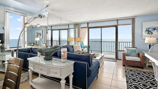 Een ruime woonkamer met een groot raam met uitzicht op zee, een witte salontafel, een blauwe bank en een rieten fauteuil.