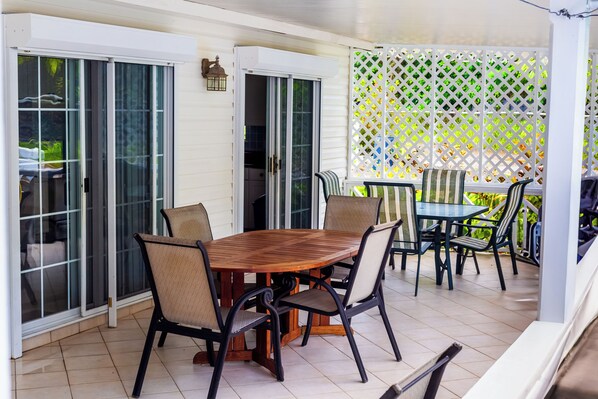 Une terrasse couverte avec une table et des chaises en bois, une petite table avec des chaises, et une clôture blanche en treillis en arrière-plan.