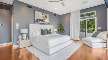A bedroom with a large bed, a nightstand, a bedside table, a ceiling fan, a window with curtains, and a view of greenery outside.