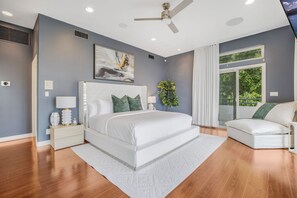 A bedroom with a large bed, a nightstand, a bedside table, a ceiling fan, a window with curtains, and a view of greenery outside.
