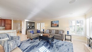 A spacious living room with a grey sofa, a patterned armchair, and a coffee table. There is a kitchen area with wooden cabinets and a dining table. - Luxurious 4 Bedroom with many Updates! (Hilton Head)