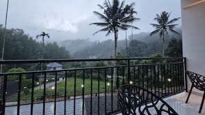 A balcony with a view of a lush landscape, including palm trees and a distant building. - Mount Minar (Devikolam)
