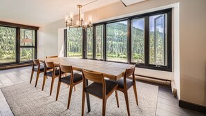 A dining area with a large table and chairs, large windows with a forest view, and a modern chandelier. - 1776 Decatur 4 Bedroom Condo (Keystone)