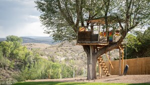 A treehouse with a wooden platform and a ladder, surrounded by a grassy area and a wooden fence. - The Beatrice Estate 7 Bedroom Estate (Grand Junction)