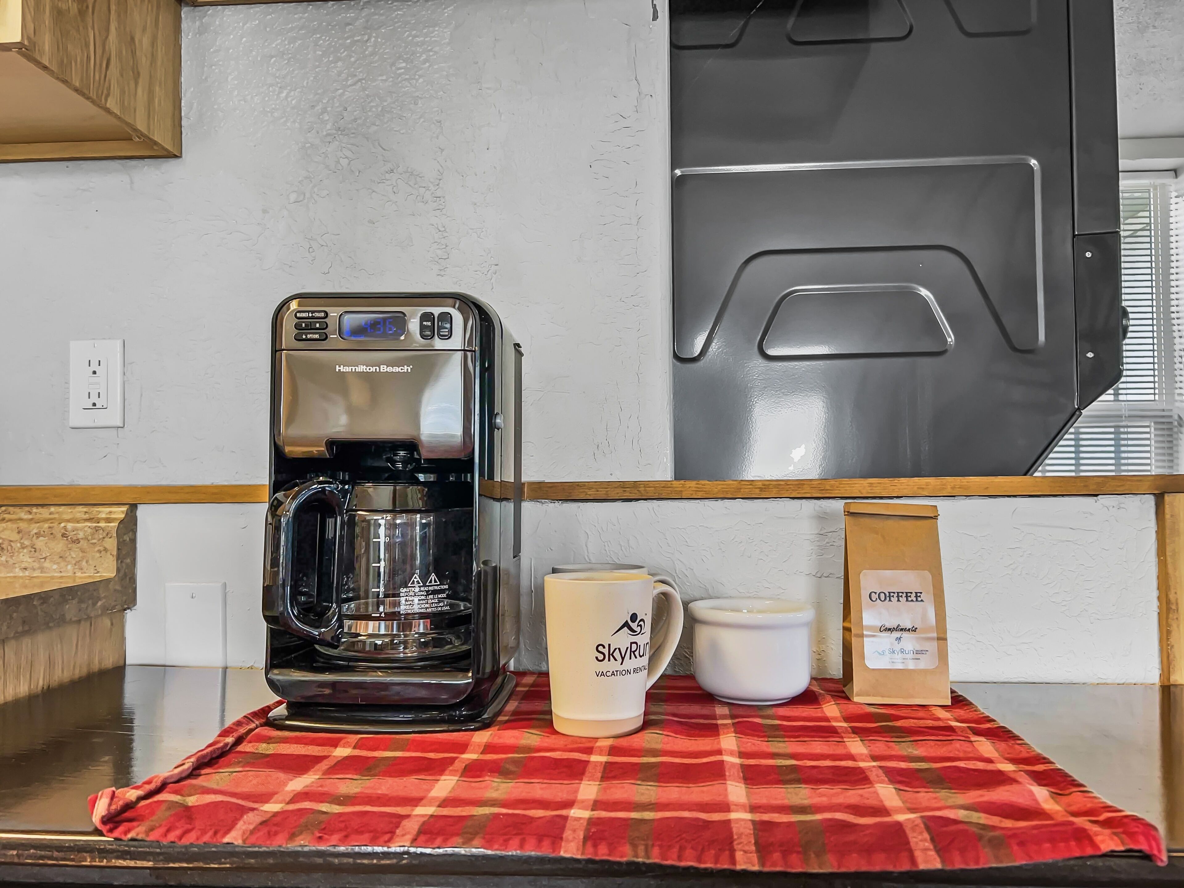 Eine Kaffeemaschine auf einer Arbeitsfläche mit einer rotkarierten Tischdecke, einer Tasse mit der Aufschrift „SkyRun Vacation Retreat“ und einer Kaffeepackung.