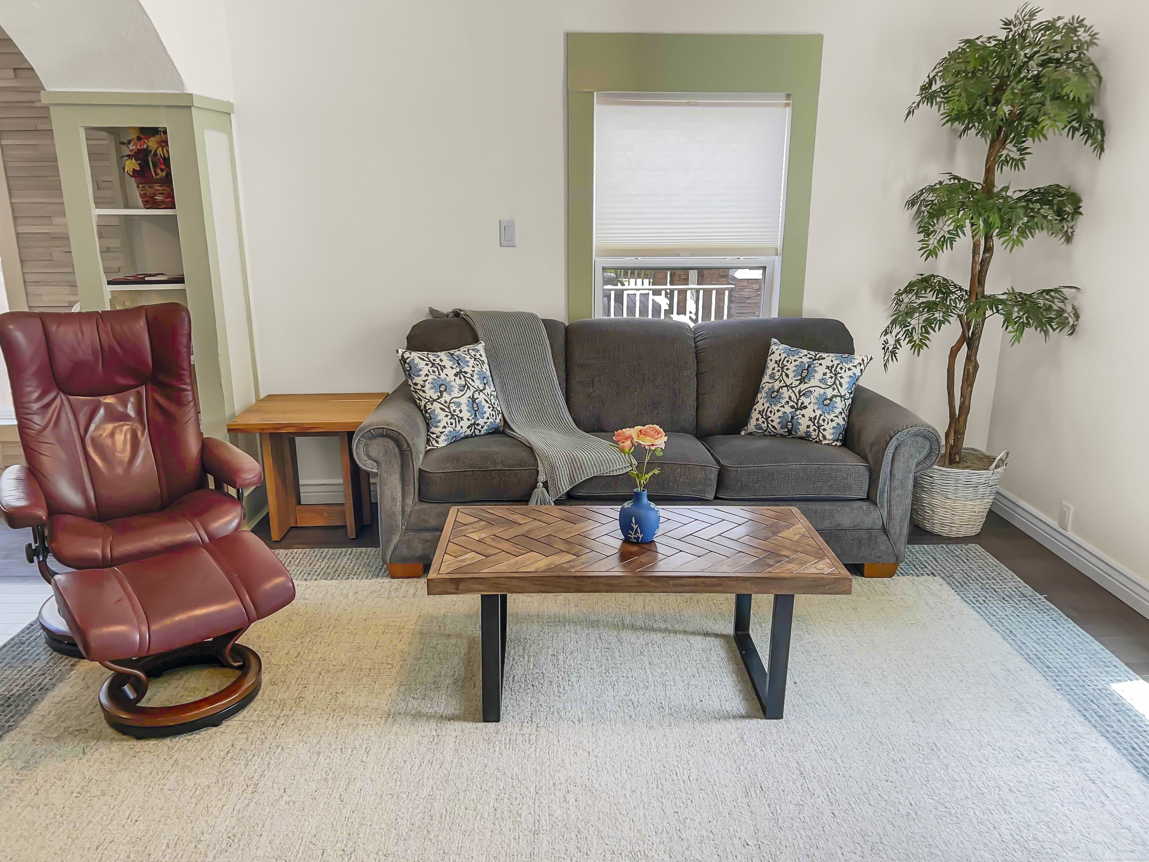 A living room with a leather chair, a sofa with patterned cushions, a wooden coffee table, and a potted plant.