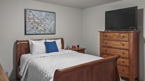 A bedroom with a wooden bed, a TV on a dresser, a nightstand with a lamp, and a painting on the wall. - Hermosa Haven 3 Bedroom Home (Montrose)