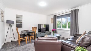 A modern living room with a brown leather sofa, a dining table with chairs, a flat-screen TV, and a large window with curtains. - Prime Commuters Choice Award by Three Bridges Apartments (Pitreavie Castle)