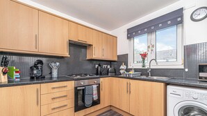 A modern kitchen with wooden cabinets, a built-in oven, a washing machine, and a window with a view of the sky. - Prime Commuters Choice Award by Three Bridges Apartments (Pitreavie Castle)
