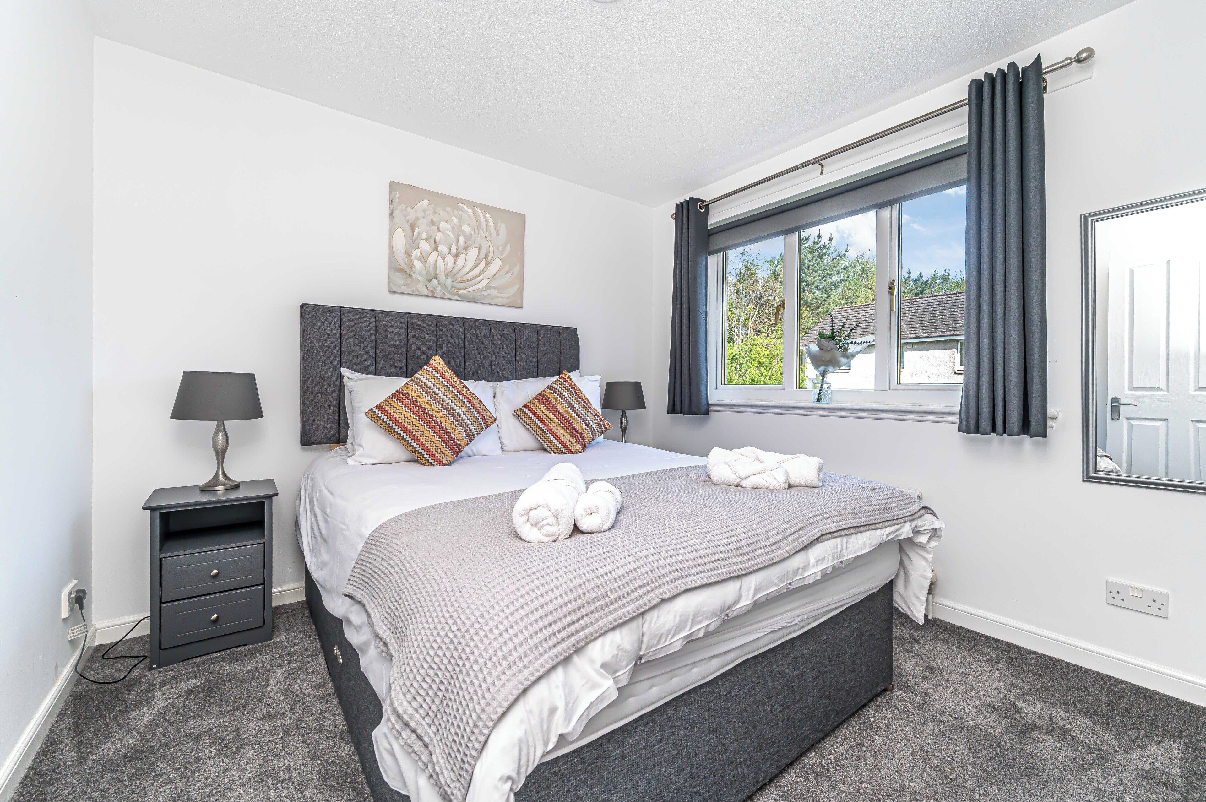 A bedroom with a bed, two bedside tables, a window with curtains, and a wall-mounted artwork.
