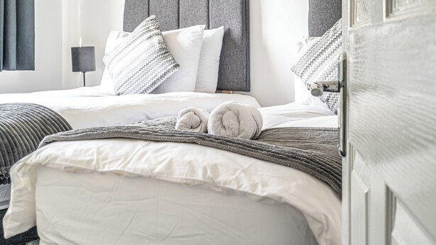 A neatly made bed with white linens and a gray headboard, a gray throw blanket, and two rolled towels on the bed. - Prime Commuters Choice Award by Three Bridges Apartments (Pitreavie Castle)