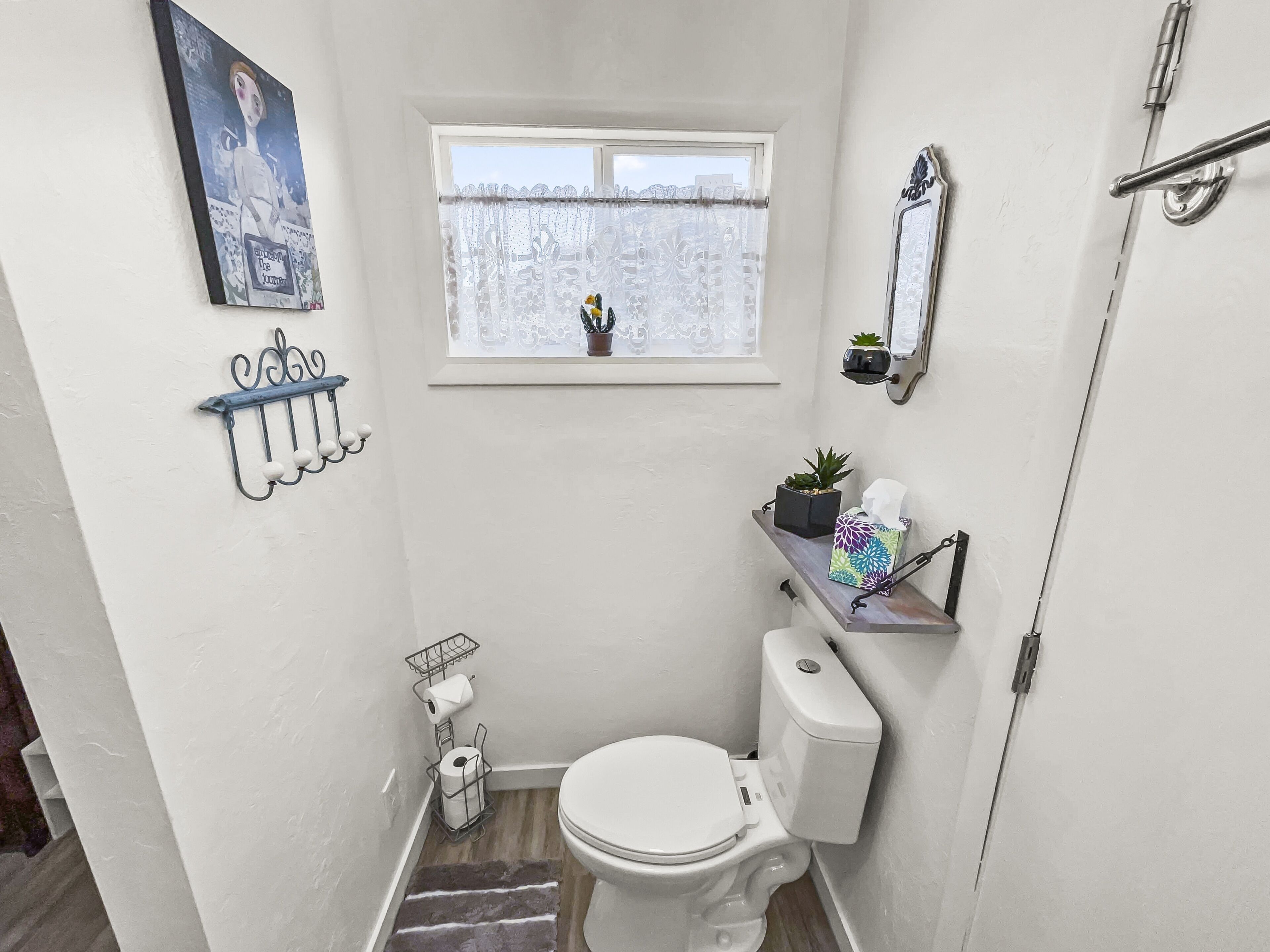 Un bagno piccolo e bianco con il water, una finestra vista, uno scaffale con una pianta e uno specchio fissato alla parete.