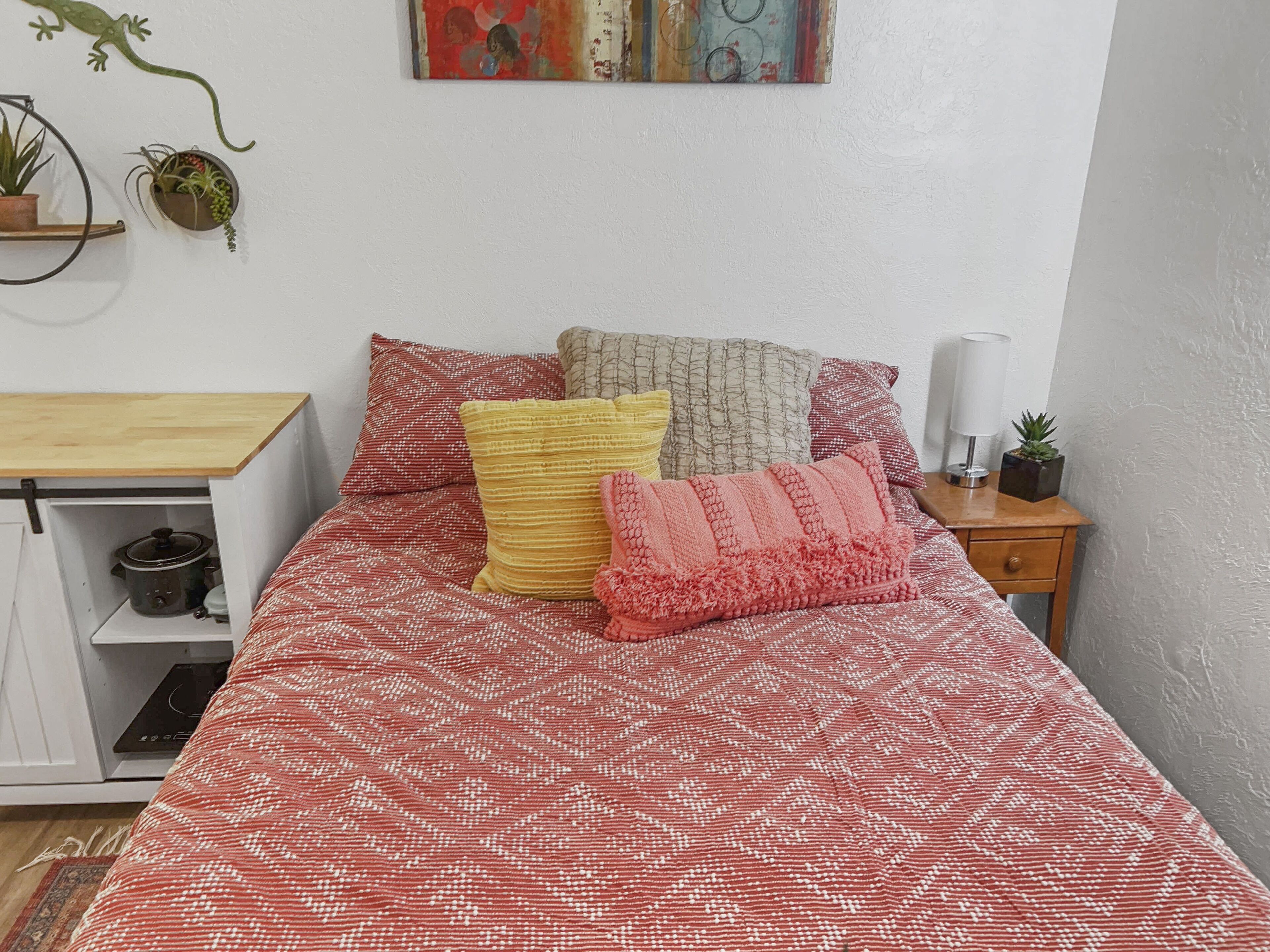 A bed with a patterned bedspread, multiple pillows, a small bedside table with a lamp, and a wall-mounted shelf with plants.
