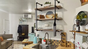 Una cucina compatta con lavandino, macchina del caffè e bollitore. Sui pensili sopra il piano di lavoro ci sono piatti e una pianta.