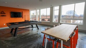 A room with a pool table, a TV mounted on the wall, and large windows offering a view of an outdoor structure. - TWO Luxe CozySuites in Downtown Cincy (Cincinnati)