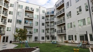 A multi-story residential building with balconies, a small lawn area with seating, and a clear sky. - TWO Luxe CozySuites in Downtown Cincy (Cincinnati)