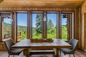 Un interno in legno con un grande tavolo e sedie, ampie finestre che si affacciano su una foresta e un lampadario.