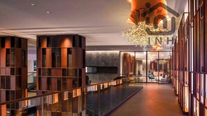 A modern hotel lobby with a glass railing, a large chandelier, and a seating area with chairs and tables.