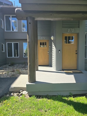 A two-story building with a porch, two doors labeled 878 and 879, and a small garden area with rocks and grass. - Snowcreek Resort Vacation Rental 879 (Mammoth Lakes)