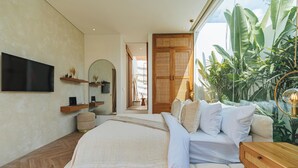 A modern bedroom with a large bed, a TV mounted on the wall, a wooden floor, and a large window with plants.