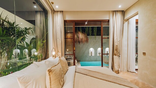 A bedroom with a large bed, a glass wall with plants, and a view of a swimming pool.