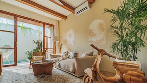 A spacious living room with a sofa, coffee table, and a wooden scooter. There is a large potted plant and a window with a view of a pool.
