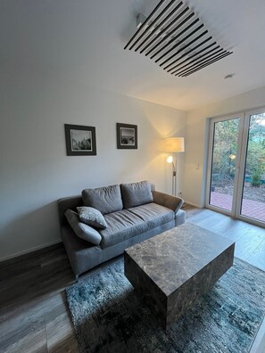 Ein modernes Wohnzimmer mit einem grauen Sofa, einem Couchtisch und einer Stehlampe. An der Wand hängen zwei gerahmte Bilder.