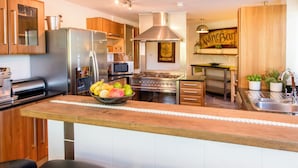A modern kitchen with wooden cabinets, stainless steel appliances, a central island with a fruit bowl, and a wine bar sign. - Large home. Close to Beach, all en-suite bedrooms. Peaceful Wooded valley views (St Agnes)