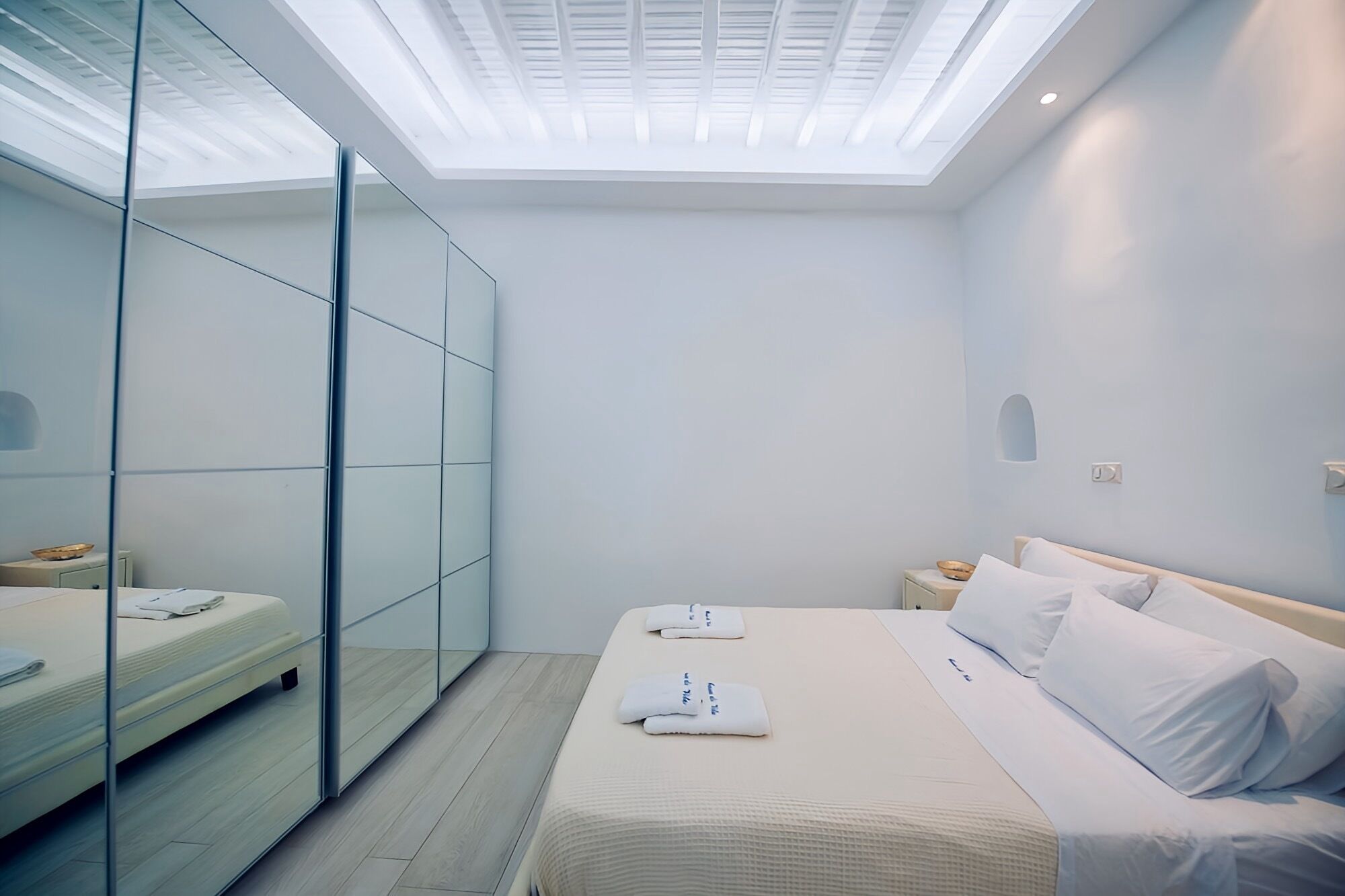 A modern bedroom with a skylight, a bed with white linens, a bedside table, and a glass-enclosed wardrobe.