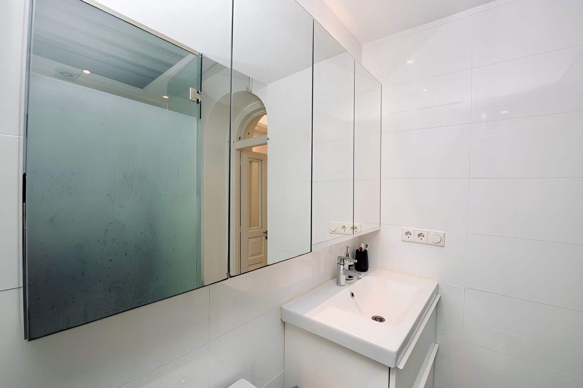 A modern bathroom with a large mirror, white tiles, a sink, and a door leading to another room.
