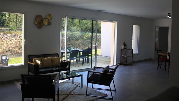 A spacious living area with a sofa, chairs, and a glass coffee table. Large windows provide a view of the outdoors.