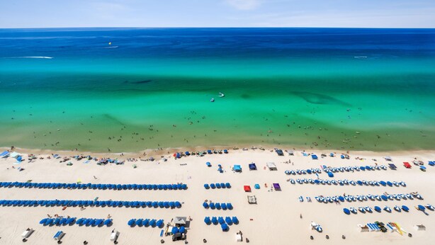 A beach with rows of blue beach chairs and umbrellas, a sandy shore, and the ocean in the background.
